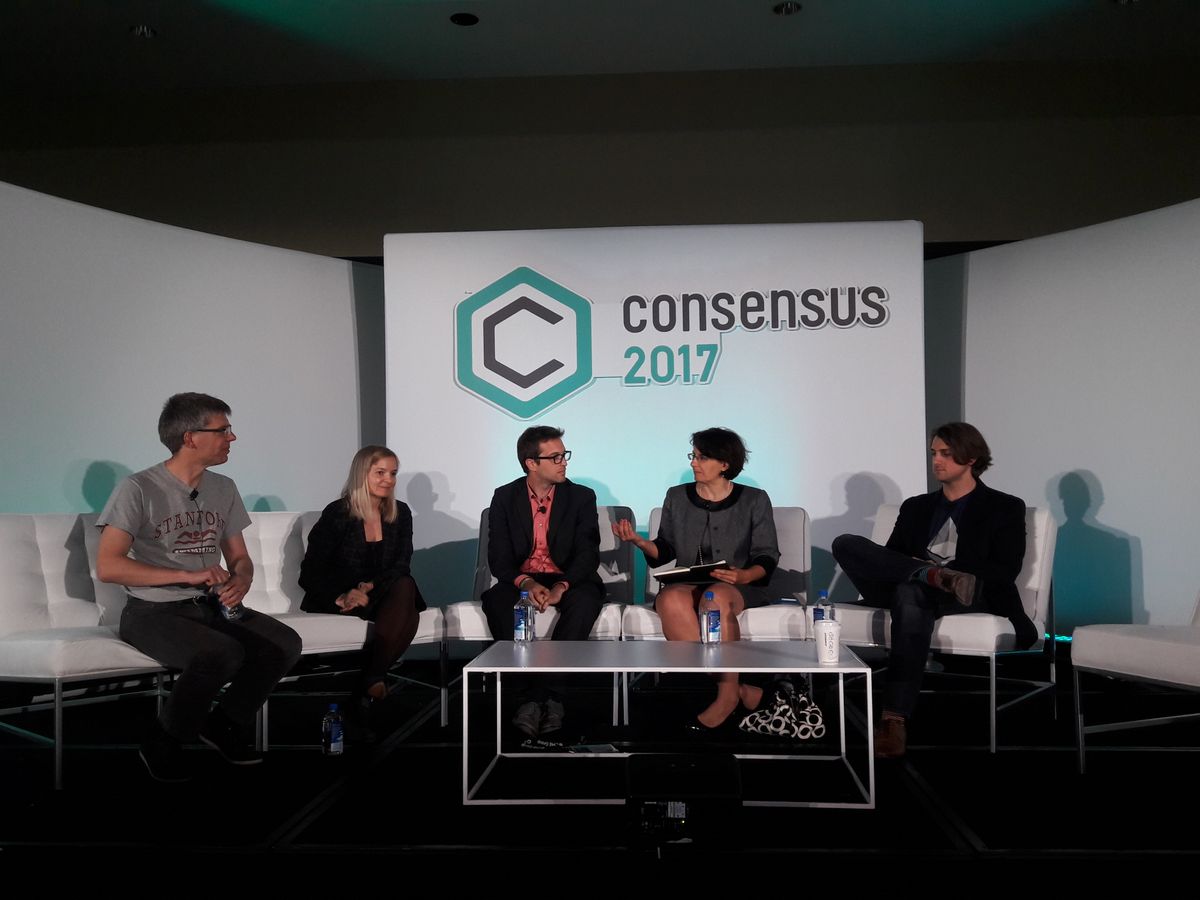 Three men and two women in white chairs around a silver coffee table. The words “Consensus 2017” are on a large white panel behind them.