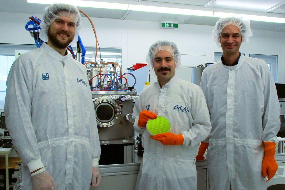 Three men in coverall suits standing in front of complicated technical equipment. The middle man holds a neon yellow disc in his orange gloved hands.