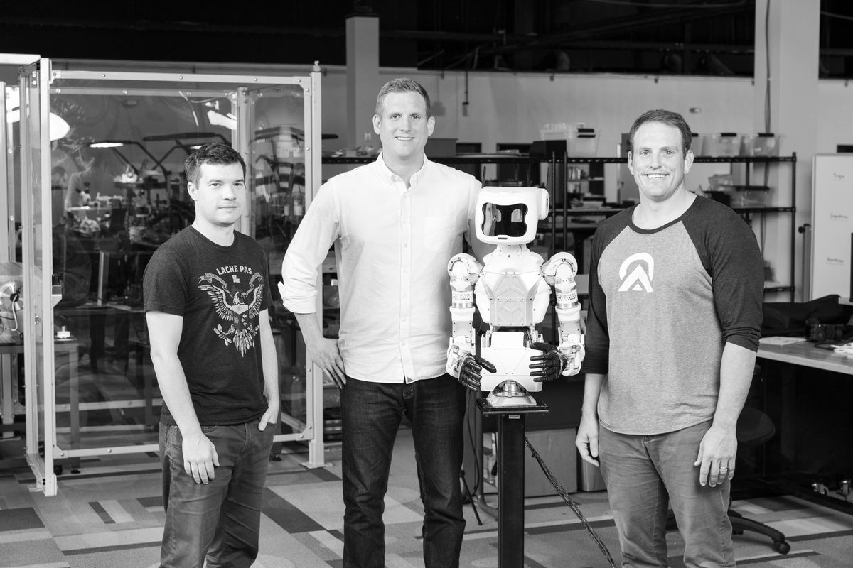 Three men stand in a robotics laboratory around a small white upper body humanoid robot on a pedestal