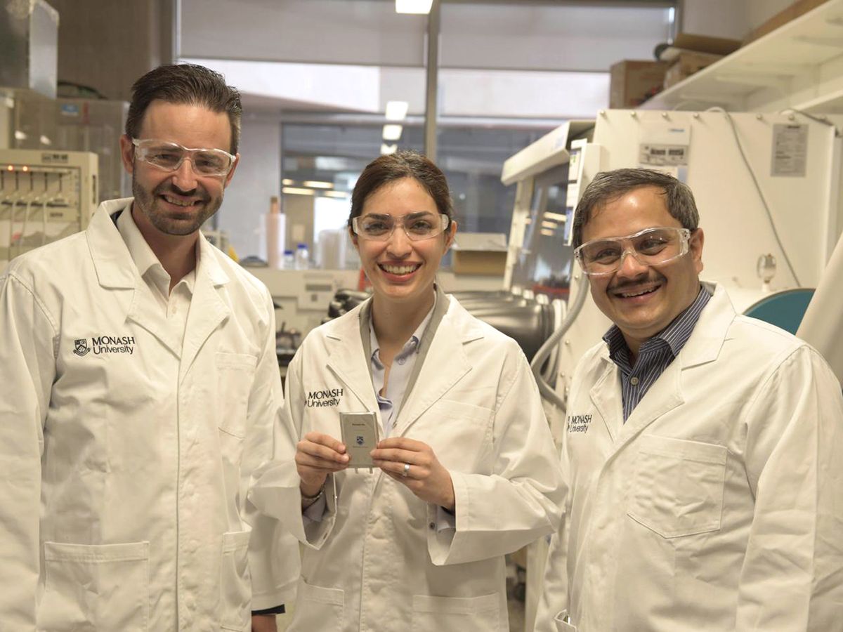 Three people in white coats and lab googles stand together in a lab while the one in the middle holds a silver object.