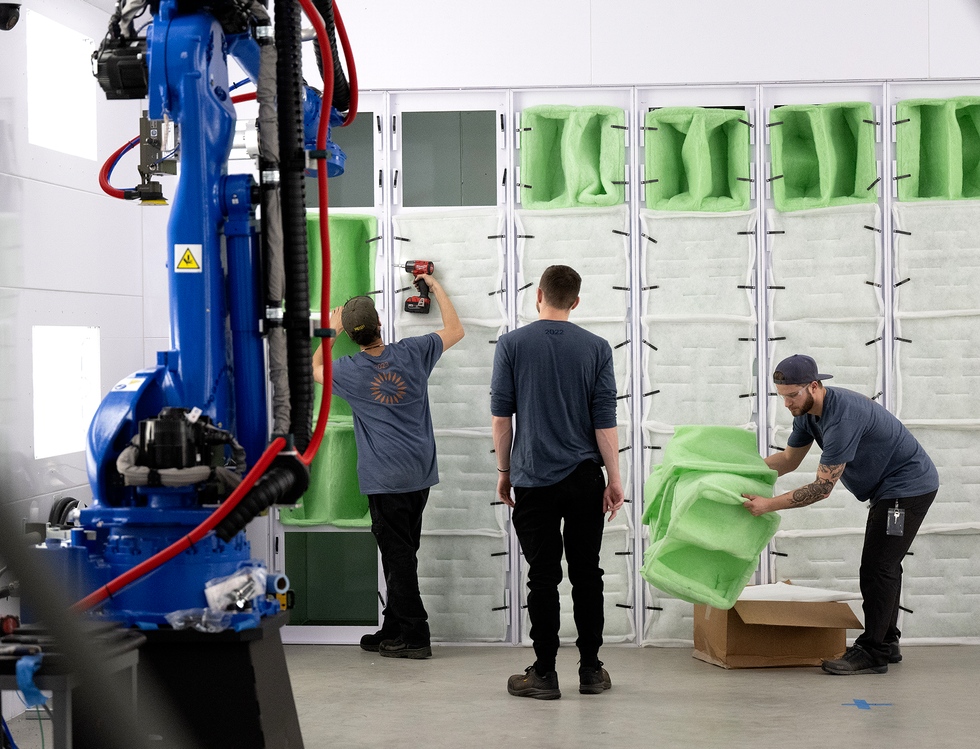 Three people insert large green pieces of foam into cube-shaped inserts in the wall behind them. In front of the people is a complicated blue robotic arm.