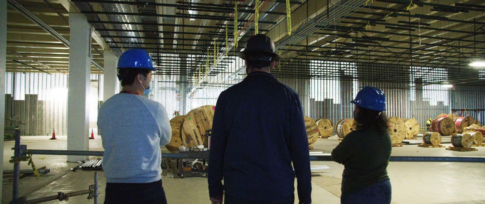 Three people wearing construction helmets in the foreground look out to an empty warehouse-sized room where giant spools of something sit.