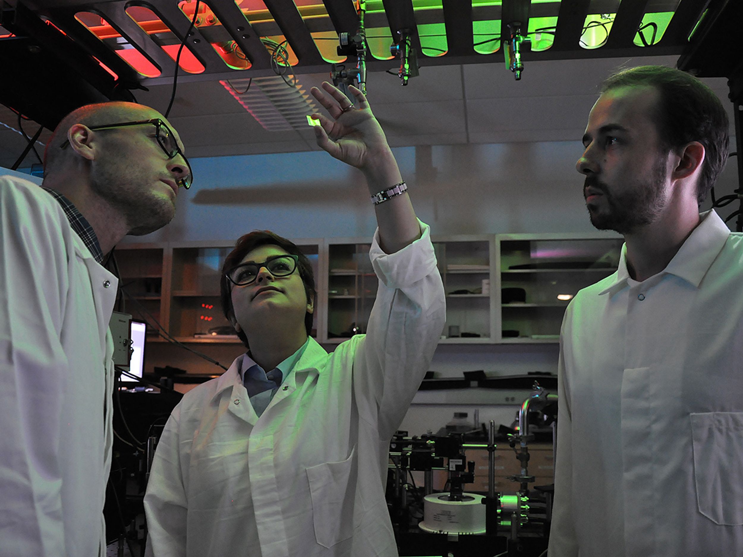Three researchers in white lab coats look at a tiny photodetector that one holds up under an array of colored lights. 