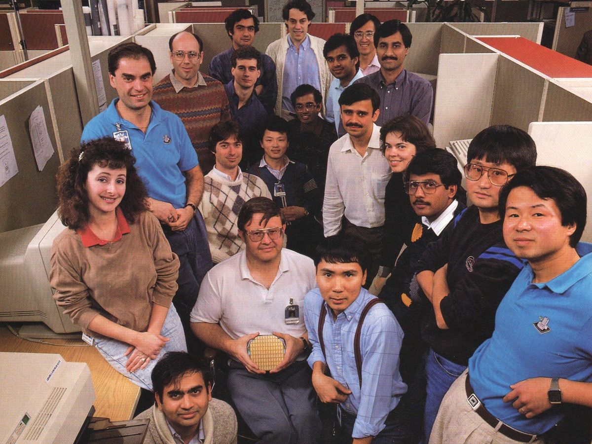 Twenty people crowd into a cubicle, the man in the center seated holding a silicon wafer full of chips
