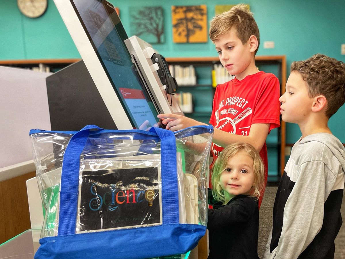 Two boys and one girl standing in front of a computer monitor. On the left side of the monitor is a backpack containing a science activity kit.
