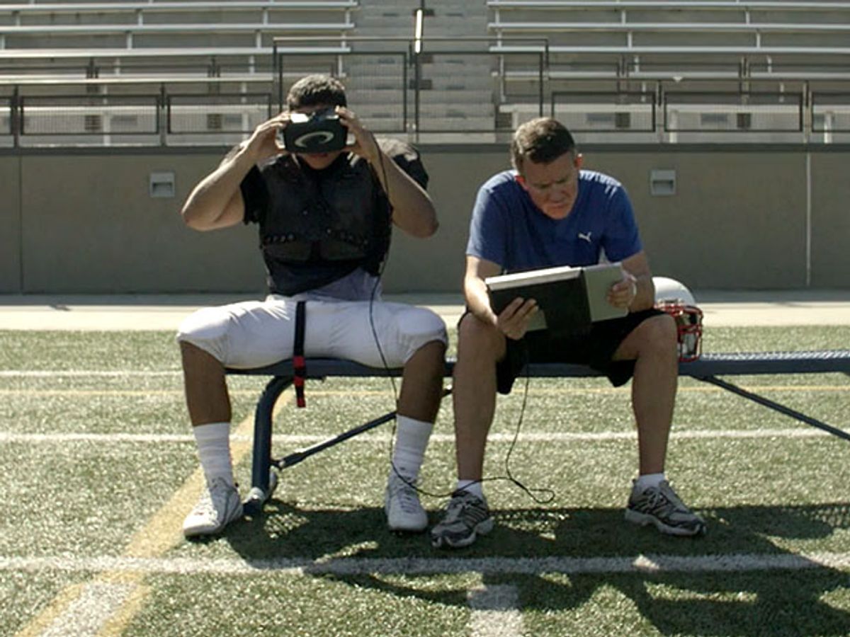 Two football players sitting next to each other on a bench, one has a VR headset on