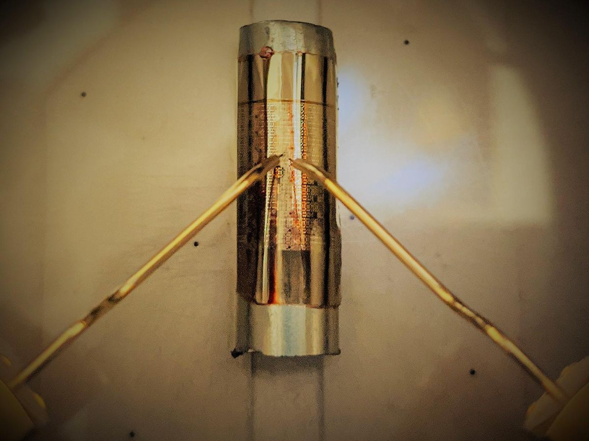 Two gold metallic probes hover over a silverish cyindrical shape, on which lies a golden shape that bends to the cylinder, and has golden and brown etchings on it.