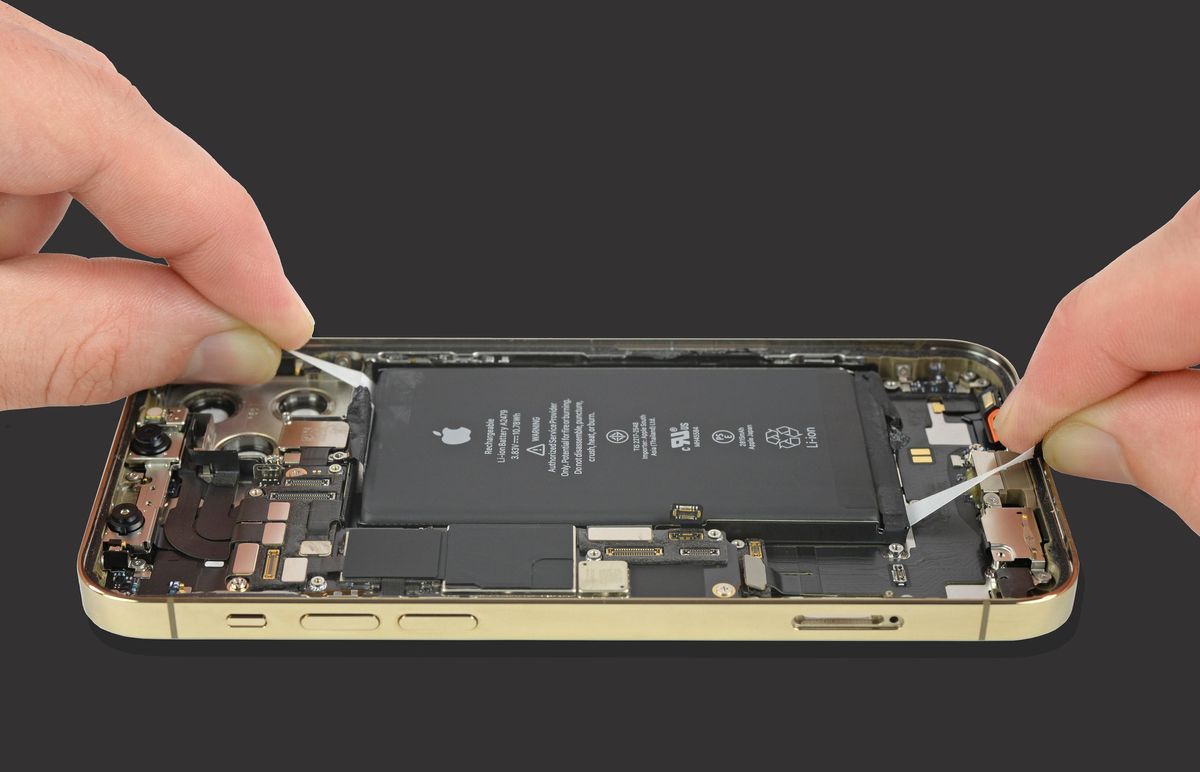 Two hands pinch adhesive beneath a large battery with an Apple icon on it in an opened phone.