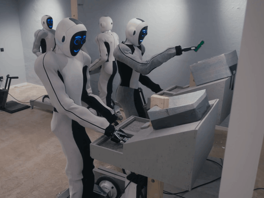 Two humanoid robots stand at a workbenches with two humanoid robots in the background