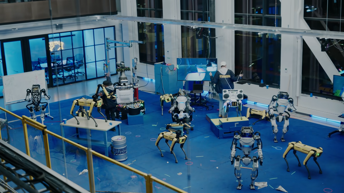 Two humans along with several Spot quadruped and Atlas humanoid robots dance in the testing area of Boston Dynamics' offices