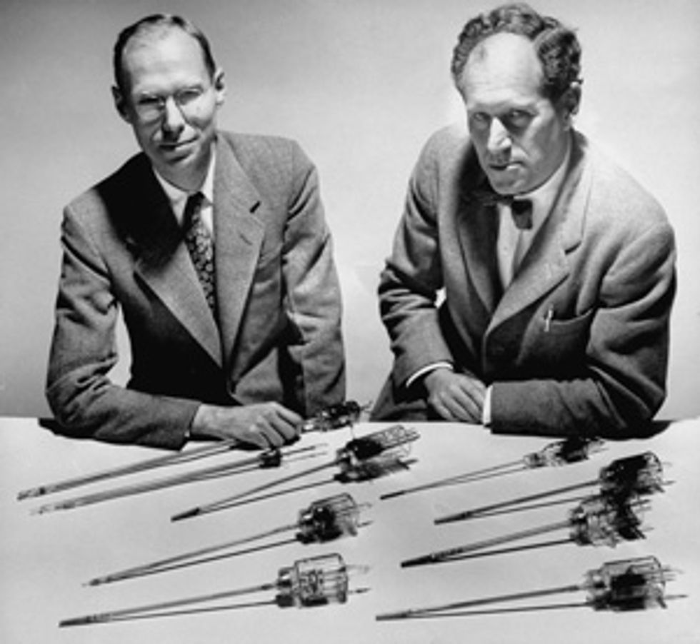 Two men behind a table with 8 different glass and metal tubes.