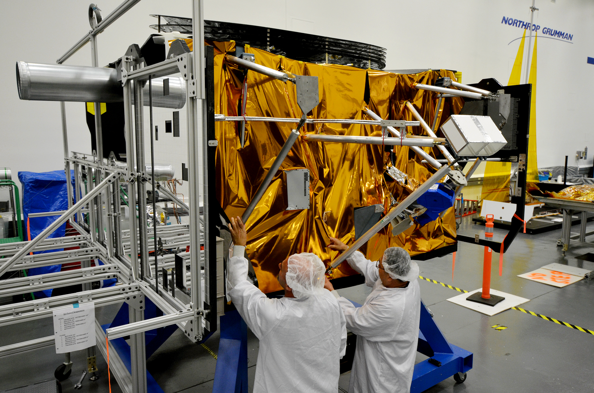Two men fix metal rods to a gold-foiled satellite component in a warehouse/clean room environment 