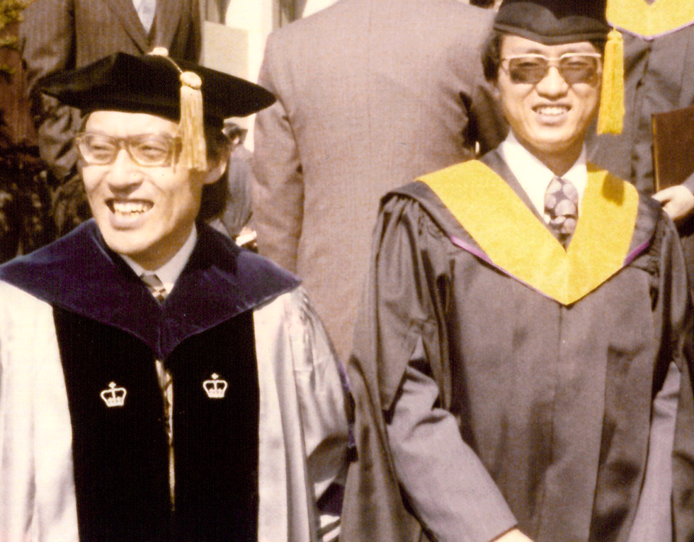 Two men in academic robes and mortar caps face the camera. Others in similar dress or in suits appear the background facing in different directions.