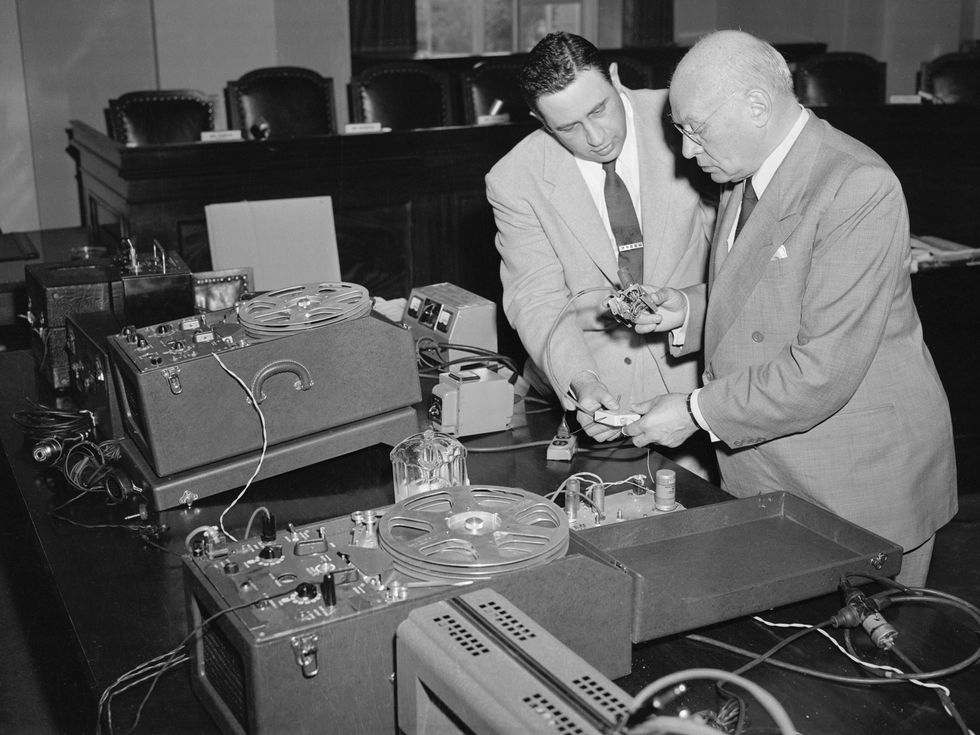 Two men in suits stand near a table heaped with tape-recording and other electronic gear.