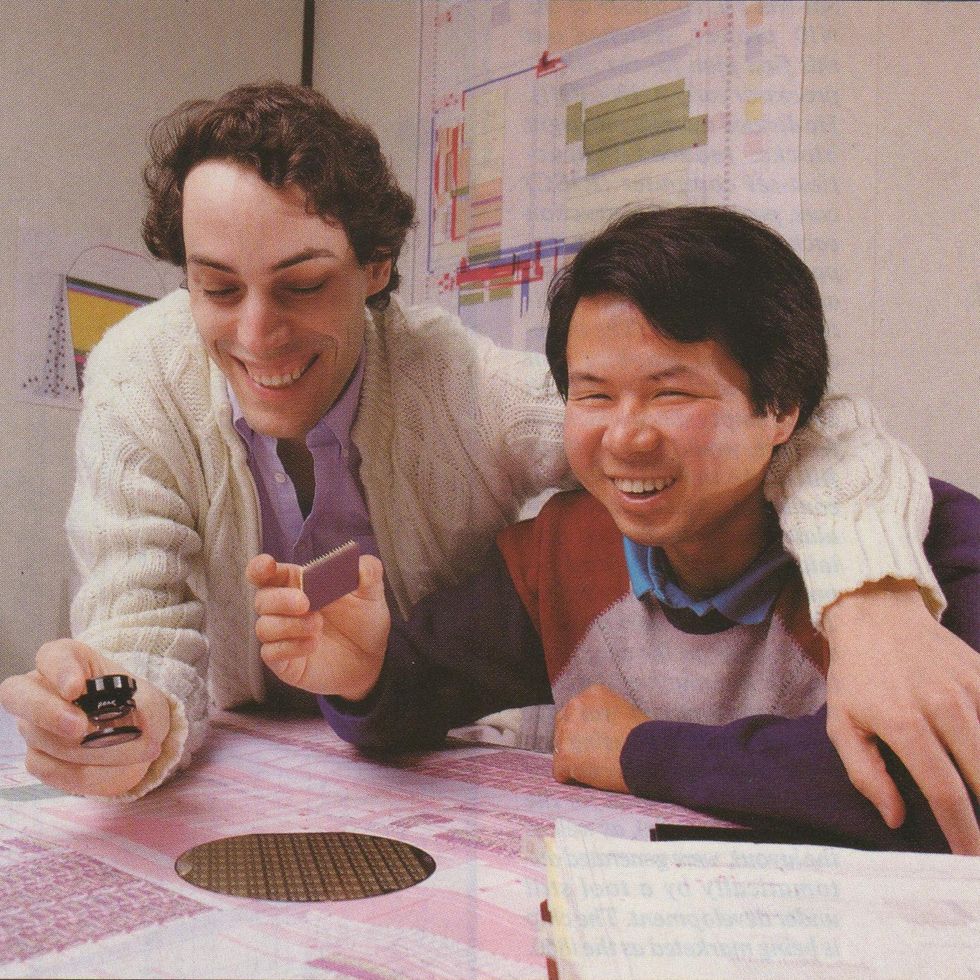 Two men lean over a circuit diagram and a silicon wafer