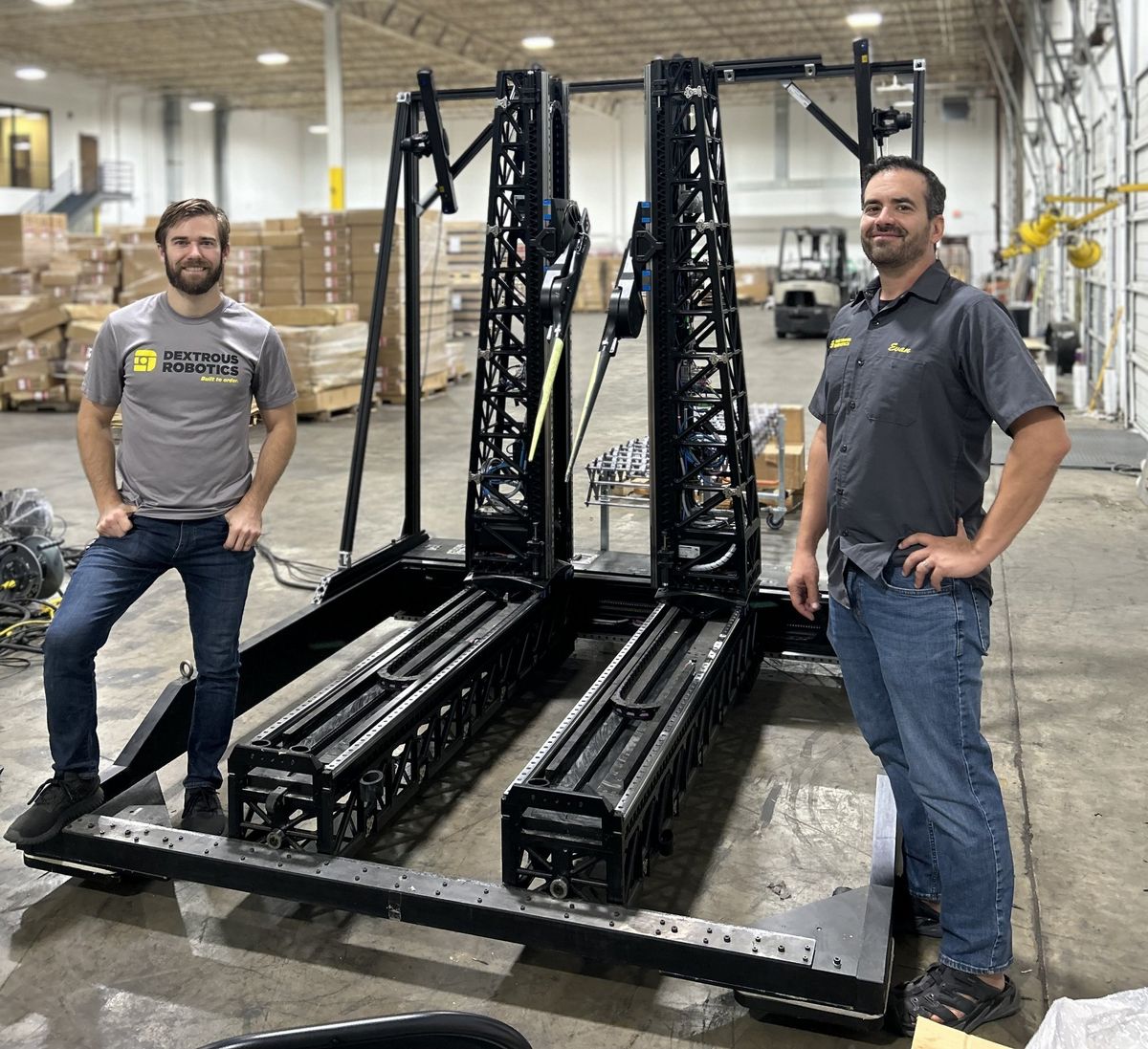 Two men stand next to a black mechanical robotic frame taller than they are, which big chopstick grippers connected to movable towers.