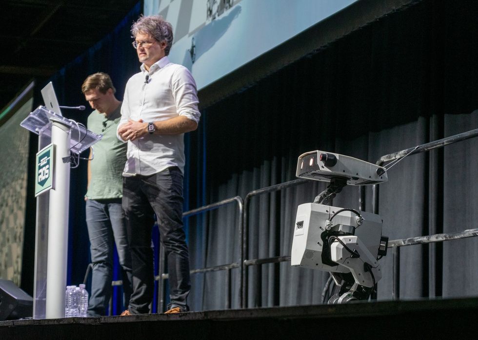 Two men stand on a conference presentation stage next to a small white legged robot.