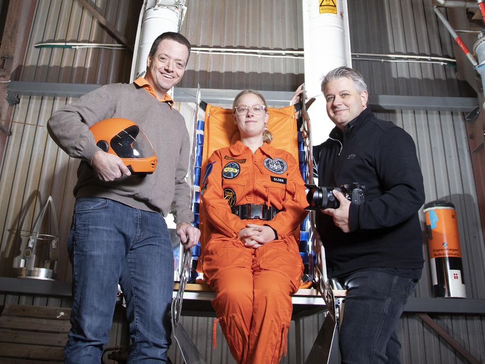 Two men stand on either side of a seated woman wearing an orange flight suit. The man on the left holds an orange flight helmet. 