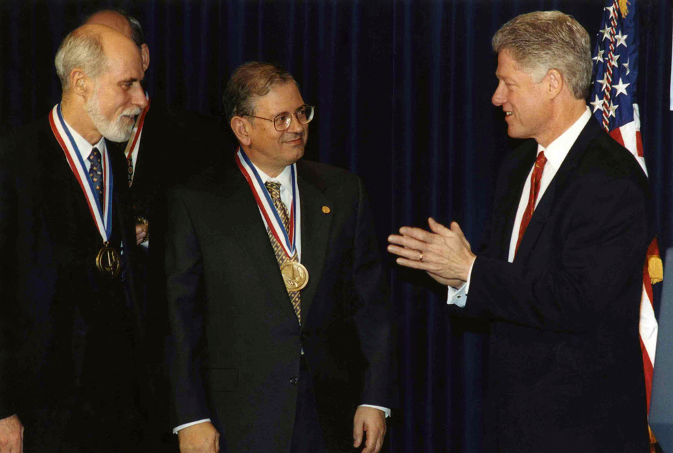 Two men wearing medals on striped ribbons around their necks chat with President Bill Clinton