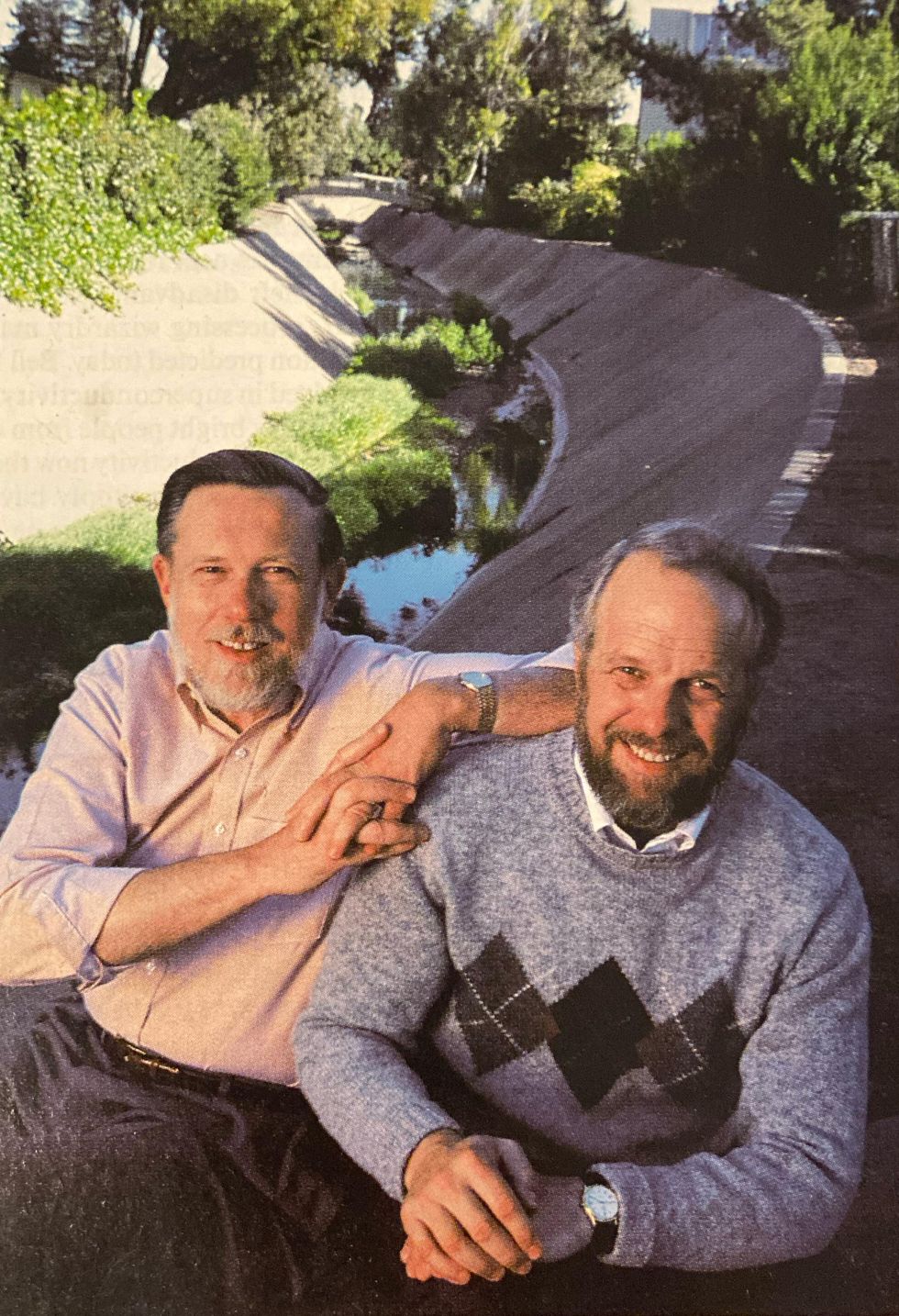 Two men with beards and mustaches seated near a creek bed with concrete sides