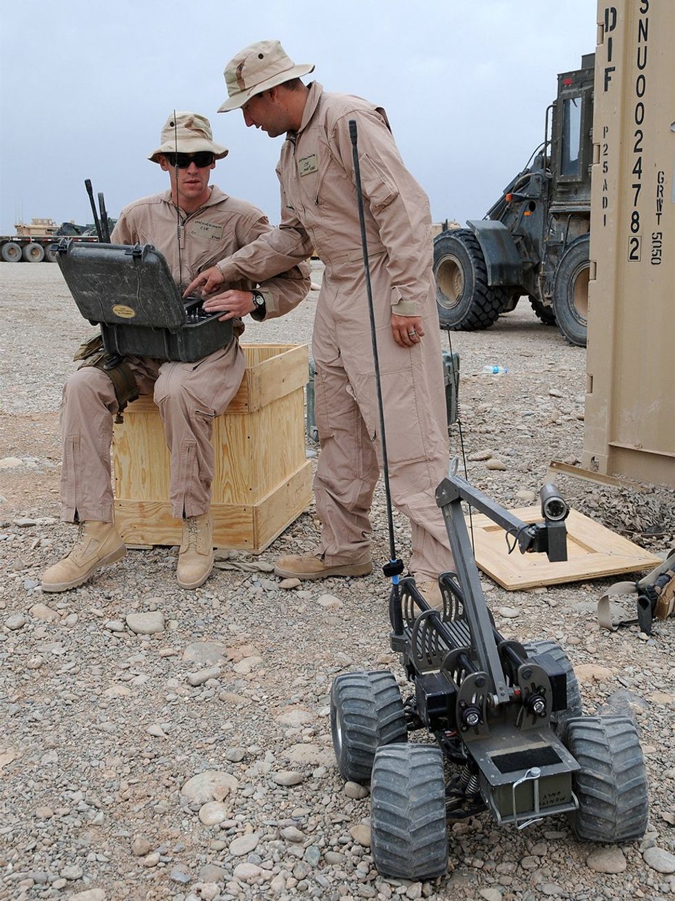 Two men work with a rugged box containing the controller for the small four-wheeled vehicle in front of them. The vehicle has a video camera mounted on a jointed arm.