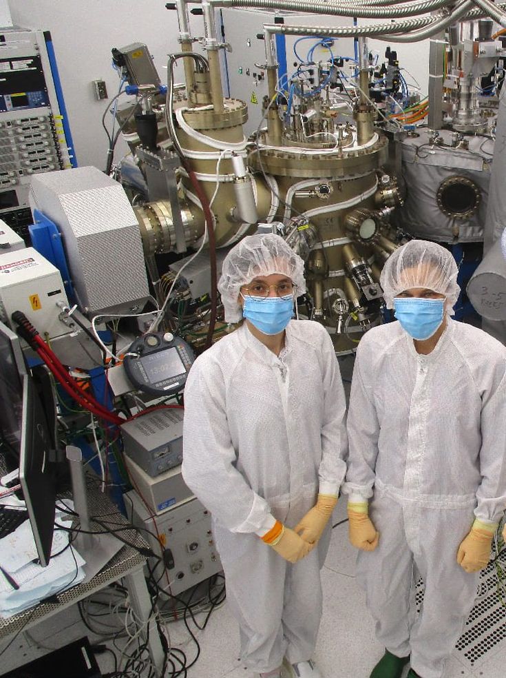Two people in clean room clothing and masks stand in front of large lab equipment.