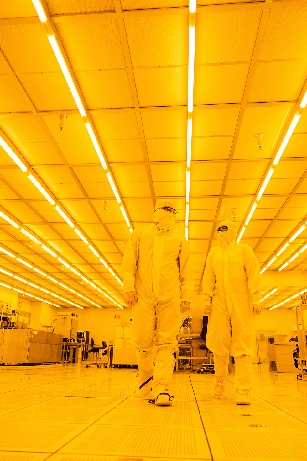 Two people, one taller than the other, walk through a yellow-lit laboratory. They are wearing white jumpsuits, face masks, and head covering.