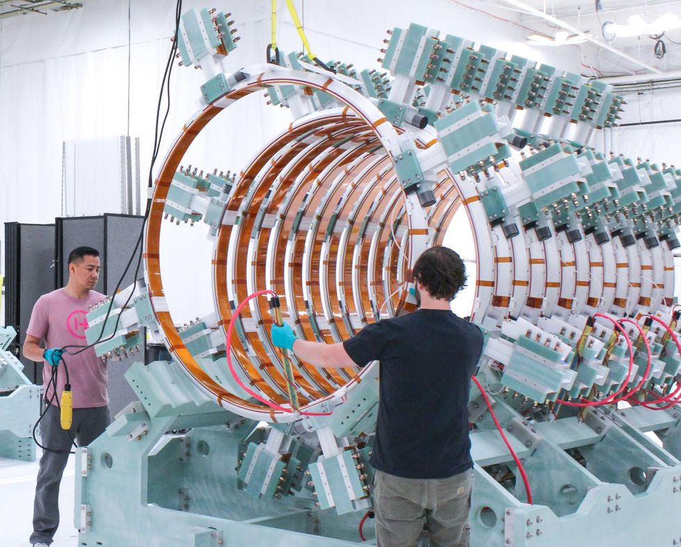 Two people stand at a larger then human device with copper coil rings in a row, and seafoam green pieces of equipment sticking out from it in a pattern of rows from six spots along the ring.