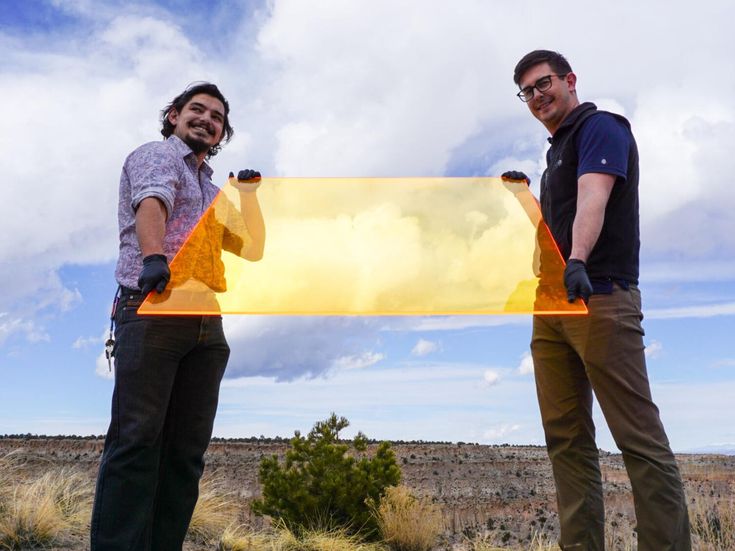 Два человека стоят в поле, держа в руках большой полупрозрачный желтый прямоугольник.