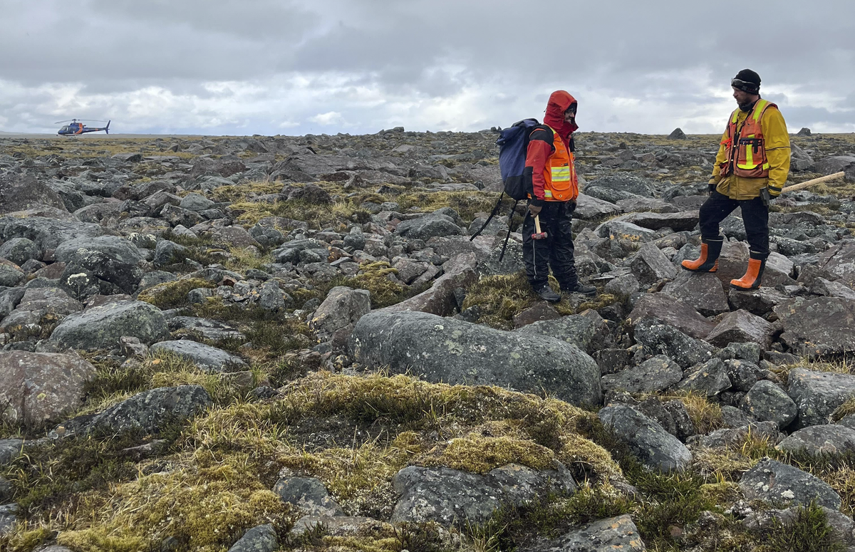 Two people stand in a gray rocky field under a cloudy sky. The person on the left is wearing a red coat, orange safety vest, and large blue backpack. The other person has a yellow jacket and orange vest.