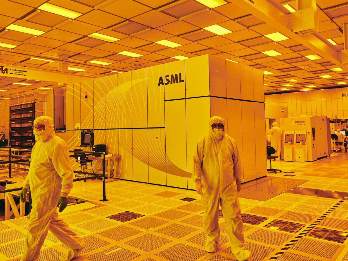 Two people walk through a room with strong yellow light. Both people are wearing full body protective suits with gloves, masks, and goggles.