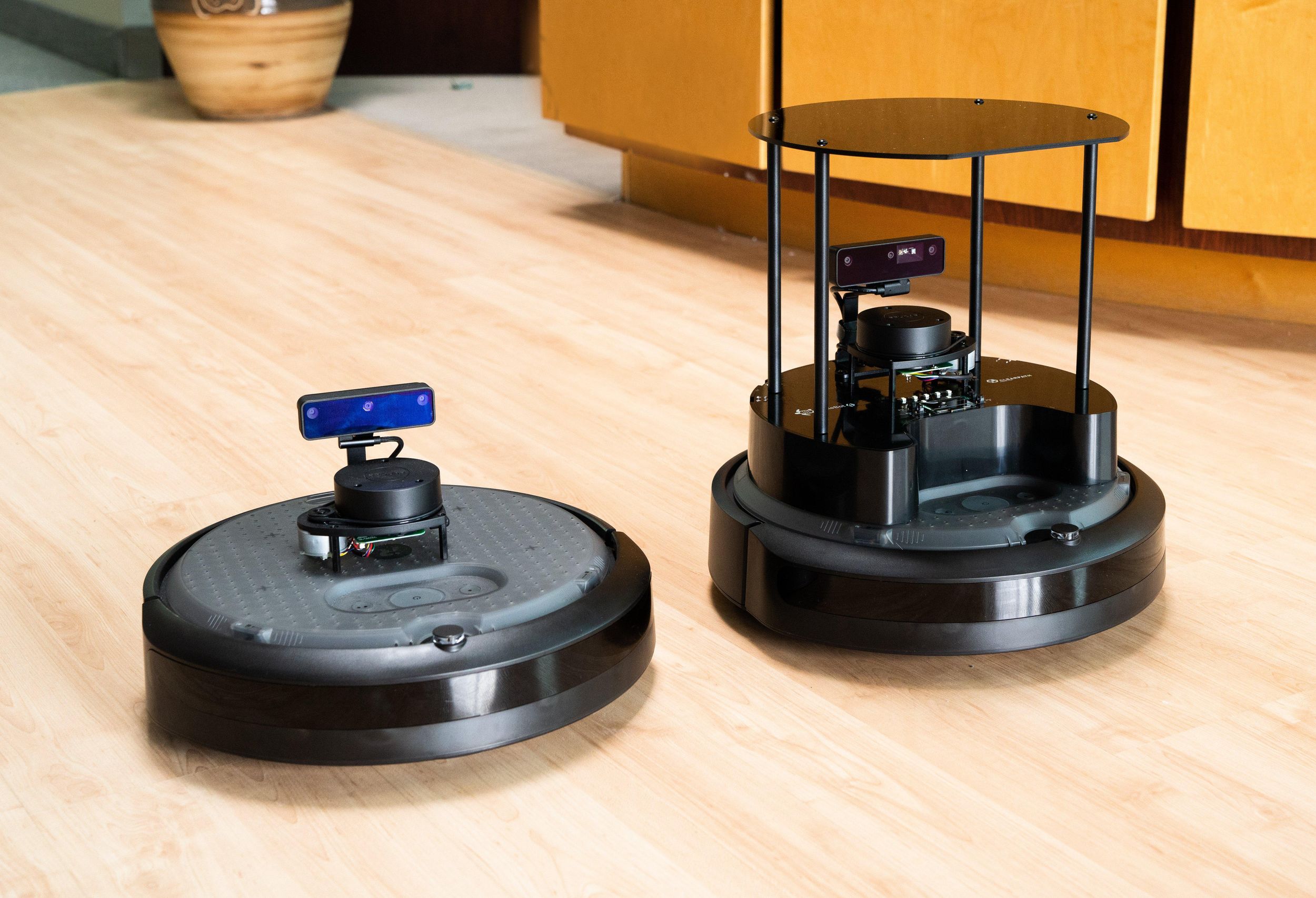 Two Roomba-like robots with sensors on top, one of which is slightly larger
