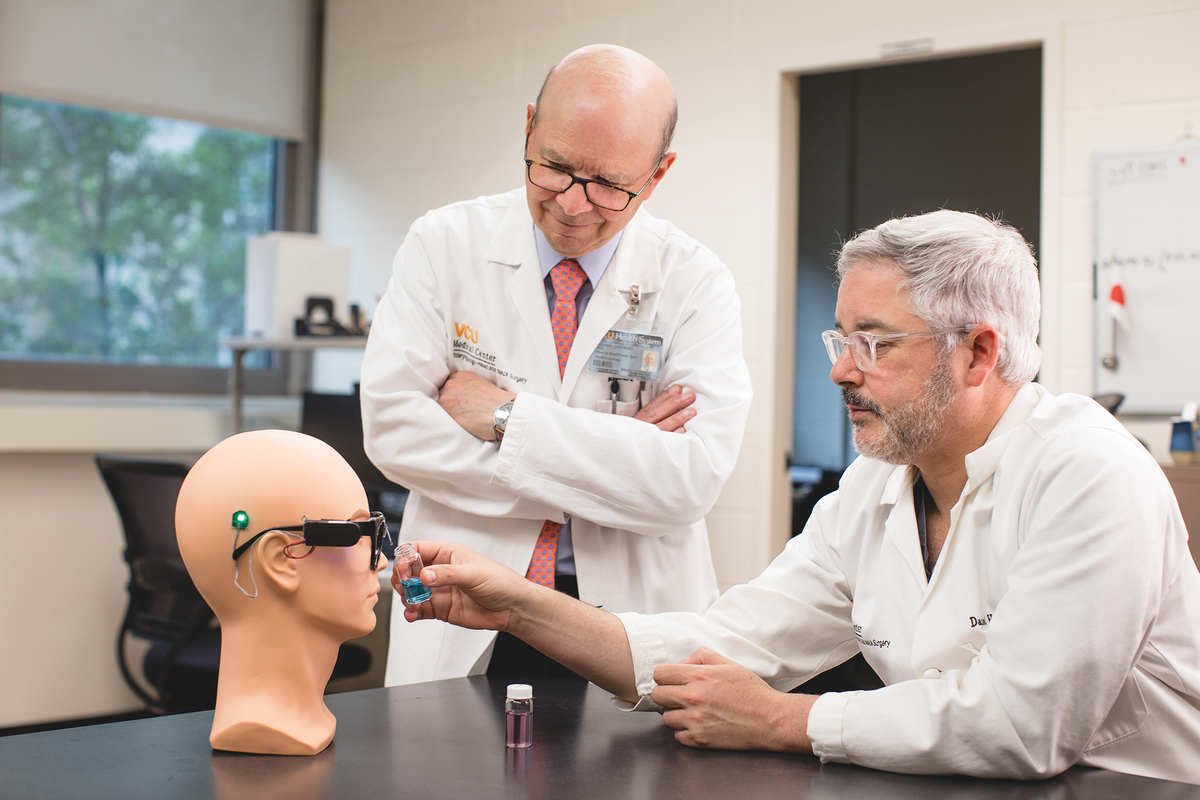 Two scientists in white lab coats look at a mannequin head that’s wearing a pair of black glasses with wires sticking out from several places. One of the scientists holds up a small vial of blue liquid to the wire above the bridge of the nose. A vial of purple liquid sits on the table. 