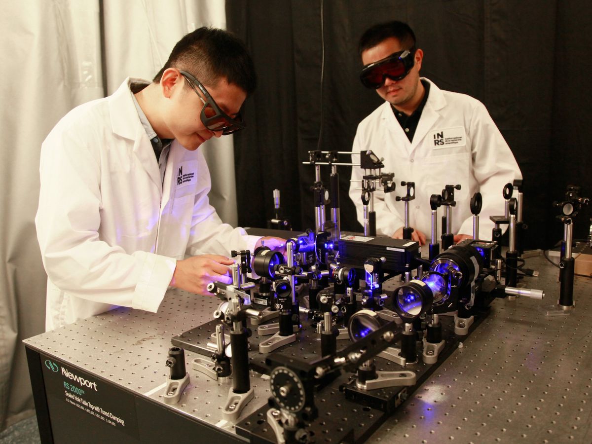 two scientists in white lab coats with goggles looking at a bunch of lenses with a purple lazer running through them