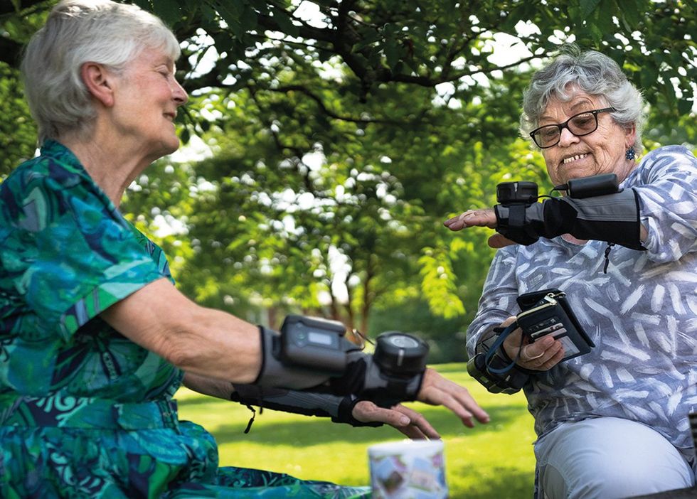 两名老年妇女各自戴着一个黑色手套状的装置，她们正在看着它。