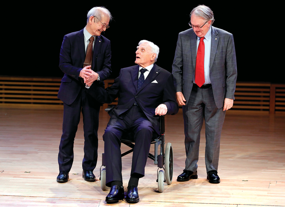 Two smiling men flank a man seated in a wheelchair, all wearing suits.