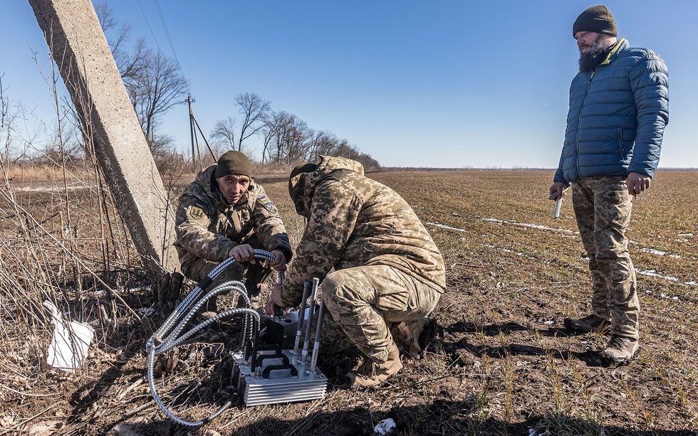 Two soldiers work on a piece of machinery consisting of a metal rectangular square with three heavy attached cables, as well as three vertical pieces coming out of it, while another man looks on.