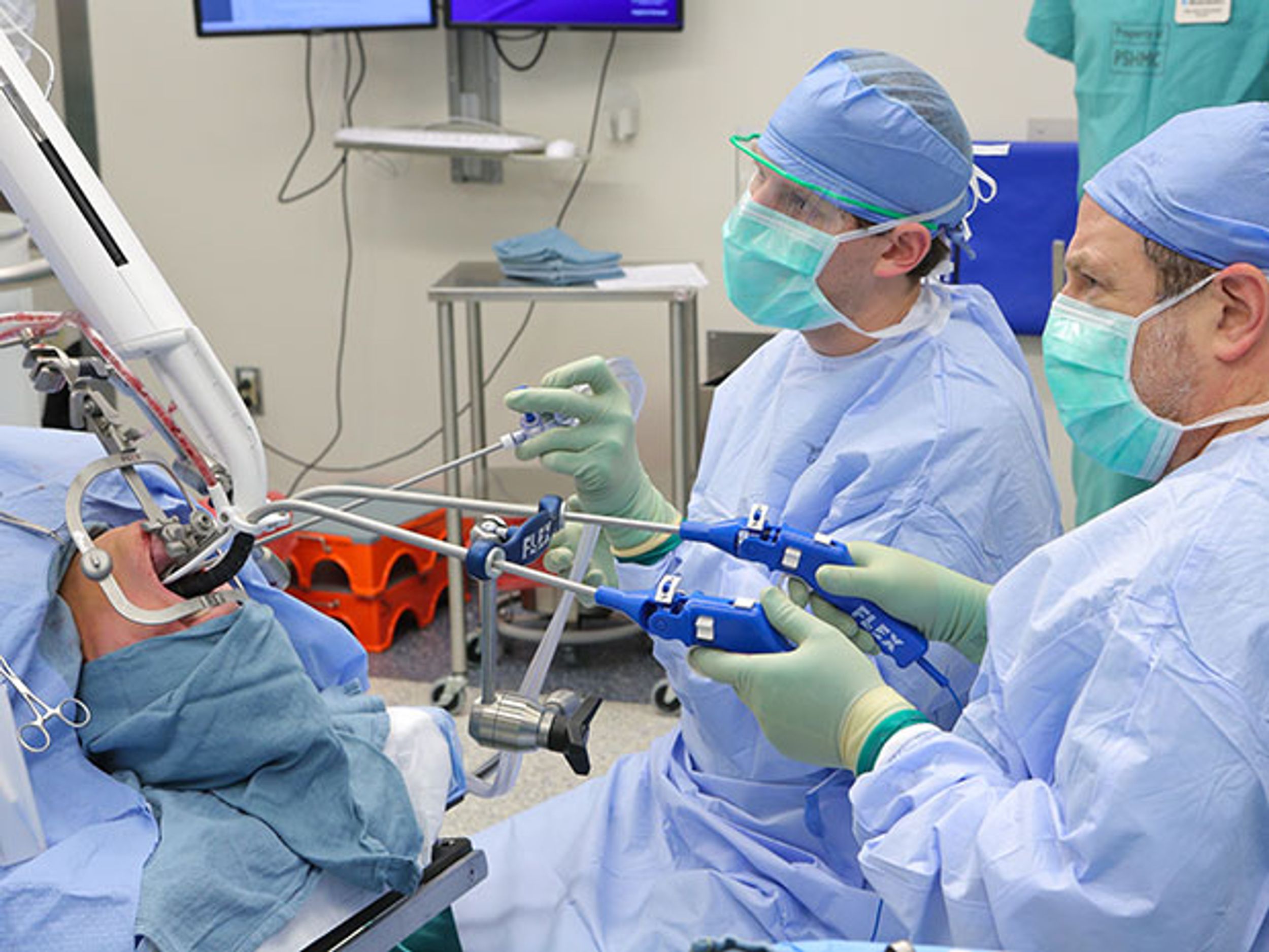 Two surgeons lean over a patient's head and insert the Flex surgical robot from Medrobotics into the patient's mouth