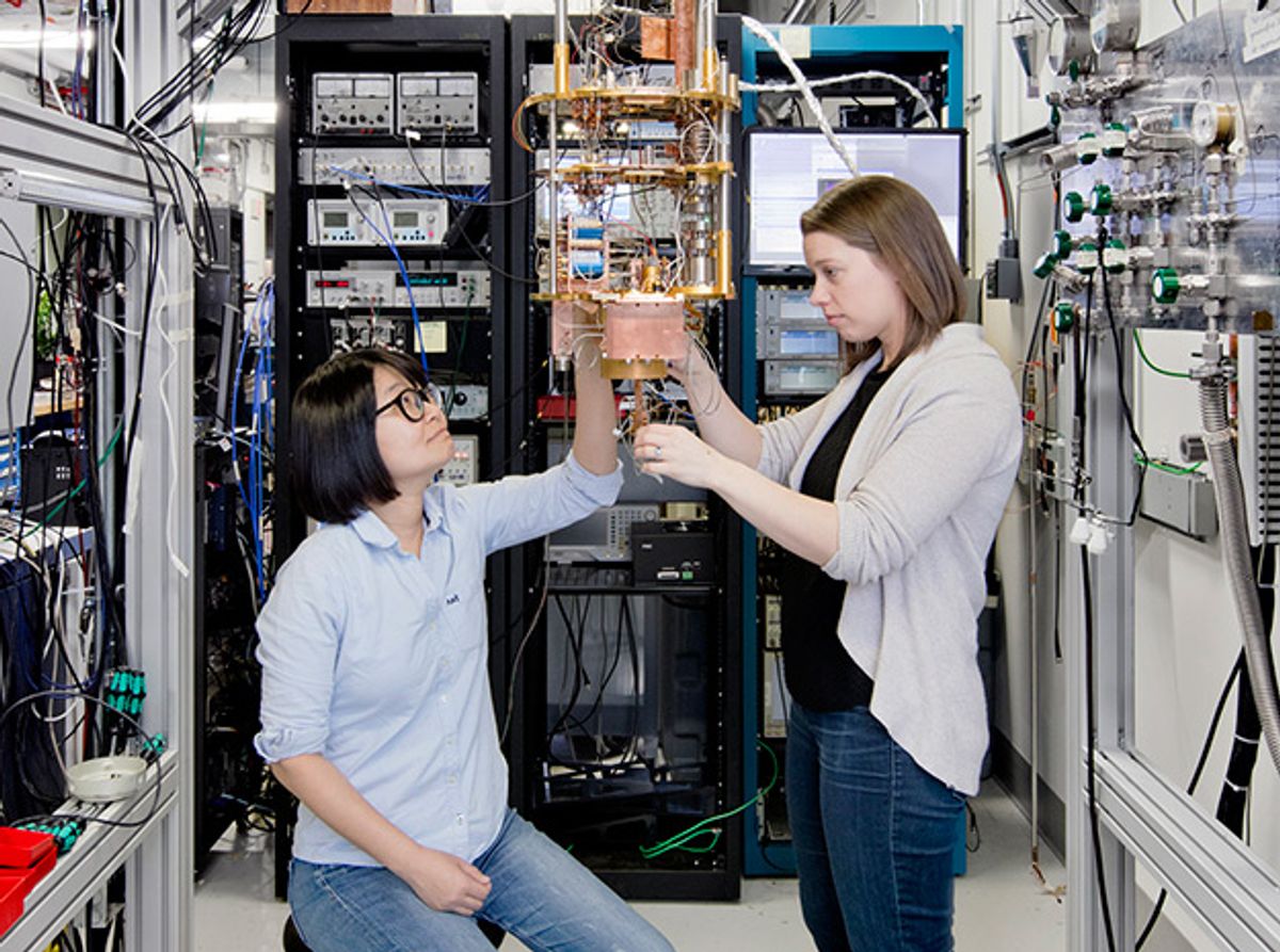 Two women surrounded by computing equipment study a device made from tubes and wires