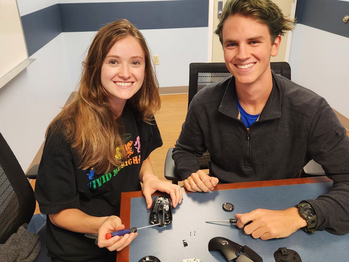 Two young students smiling for the camera with screwdrivers in their hands and a disassembled computer mouse on a table
