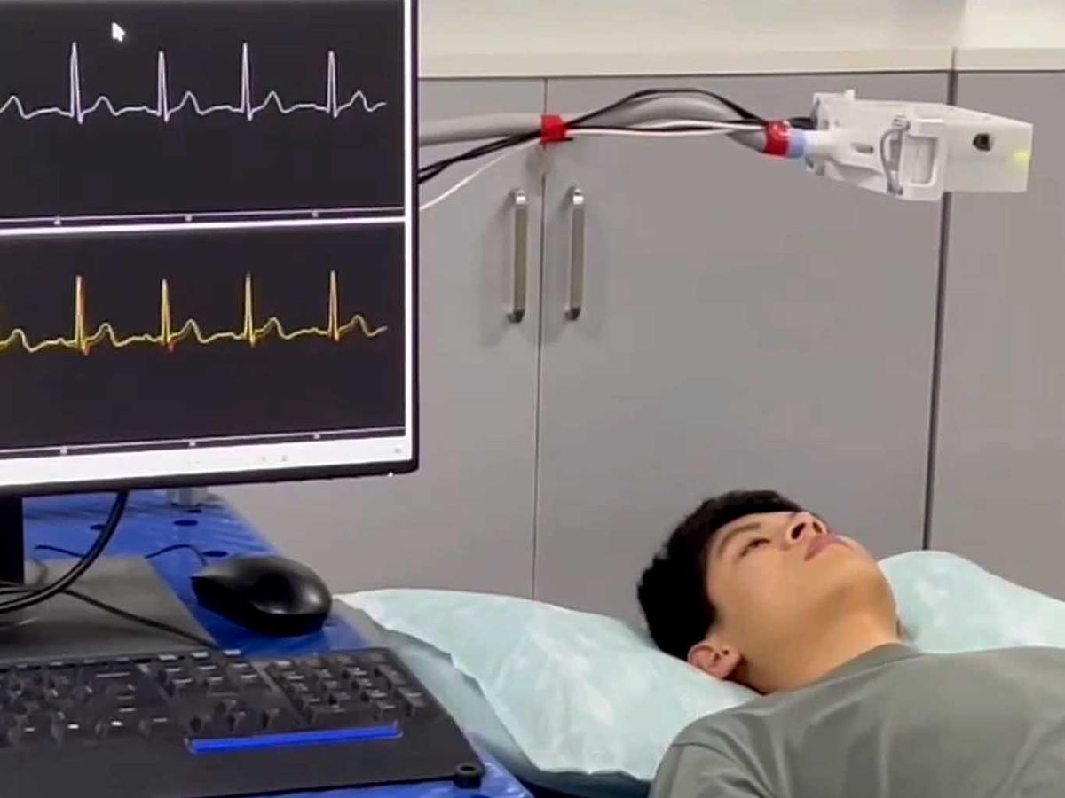 Video still of a man lying down. A box shaped device on a pole sits above his body. To the left, a monitor displays ECG readings.