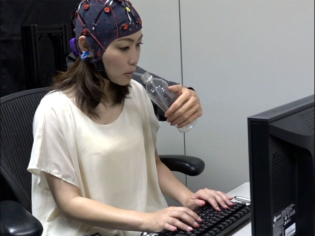 Video still showing a woman wearing the BMI electrode cap while typing with two hands and controlling the robotic 'third' arm to give her water.
