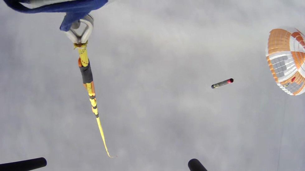  View looking down from a helicopter as it approached Electron rocket booster, descending by parachute. A yellow cable at left has a large hook at the end.