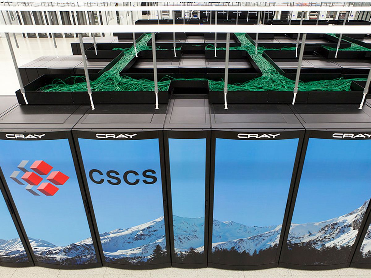  wall of cabinets show a winter mountain seen. The letters CSCS are visible on one cabinet. The word “Cray” is at the top of each cabinet. Above the cabinets a tangle of green cables extends into the distance.