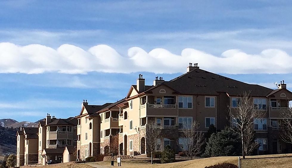 White clouds stretching from the leftmost to the rightmost upper part of a photograph form a regularly repeating wavelike pattern above and behind a block of apartments.