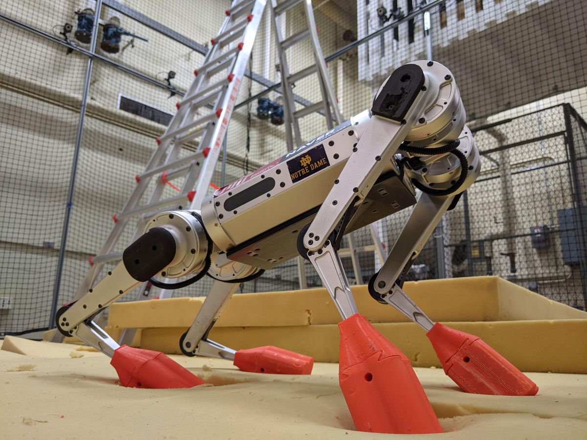 Wide angle close up picture of a silver quadrupedal robot with red 3D printed boots standing on a foam pad with a ladder in the background