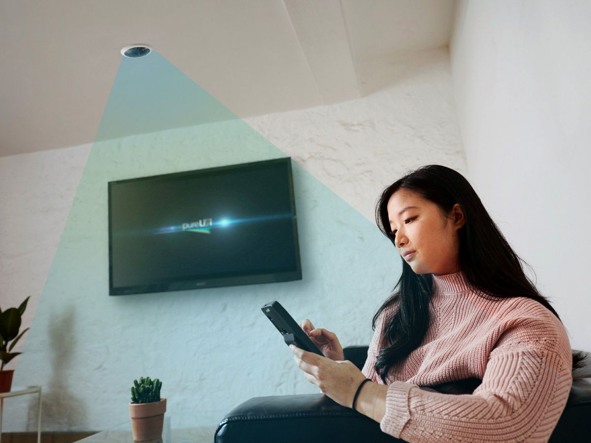 woman sitting on a couch holding a smartphone with a tv on the wall in the background and a green triangle coming out of the above light in the ceiling