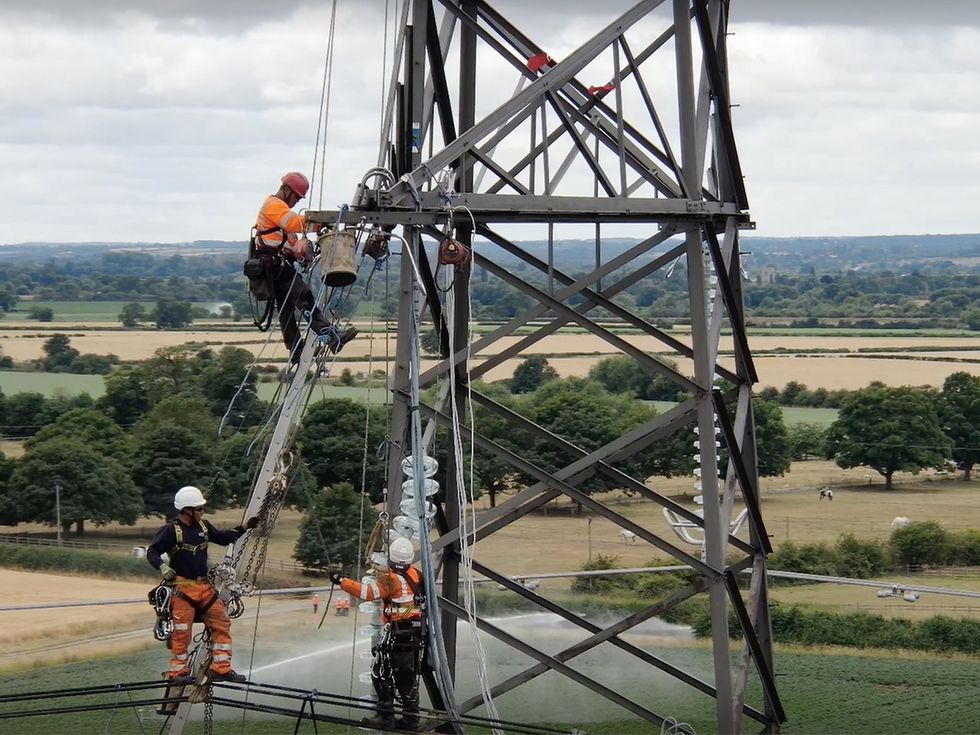 Workers in harnesses and hard hats climbing and standing high up on a transmission tower in the countryside.