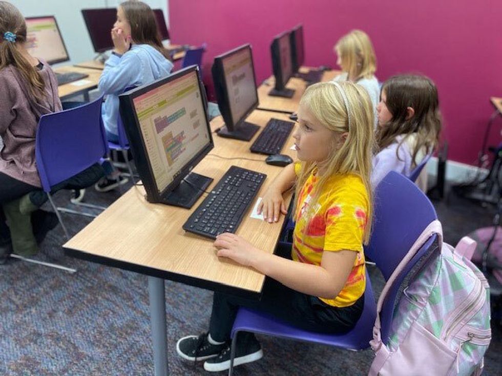 young girls sitting at a large desk with computers and keyboards in front of them, the girl closest wearing a bright yellow shirt