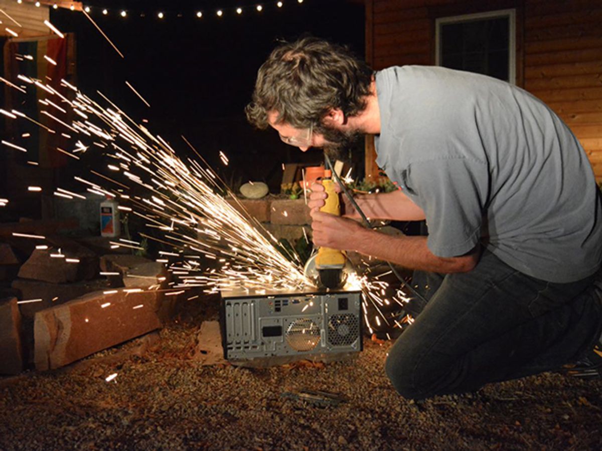 Za Wilcox on his knees destroying a computer with a power tool as sparks fly.